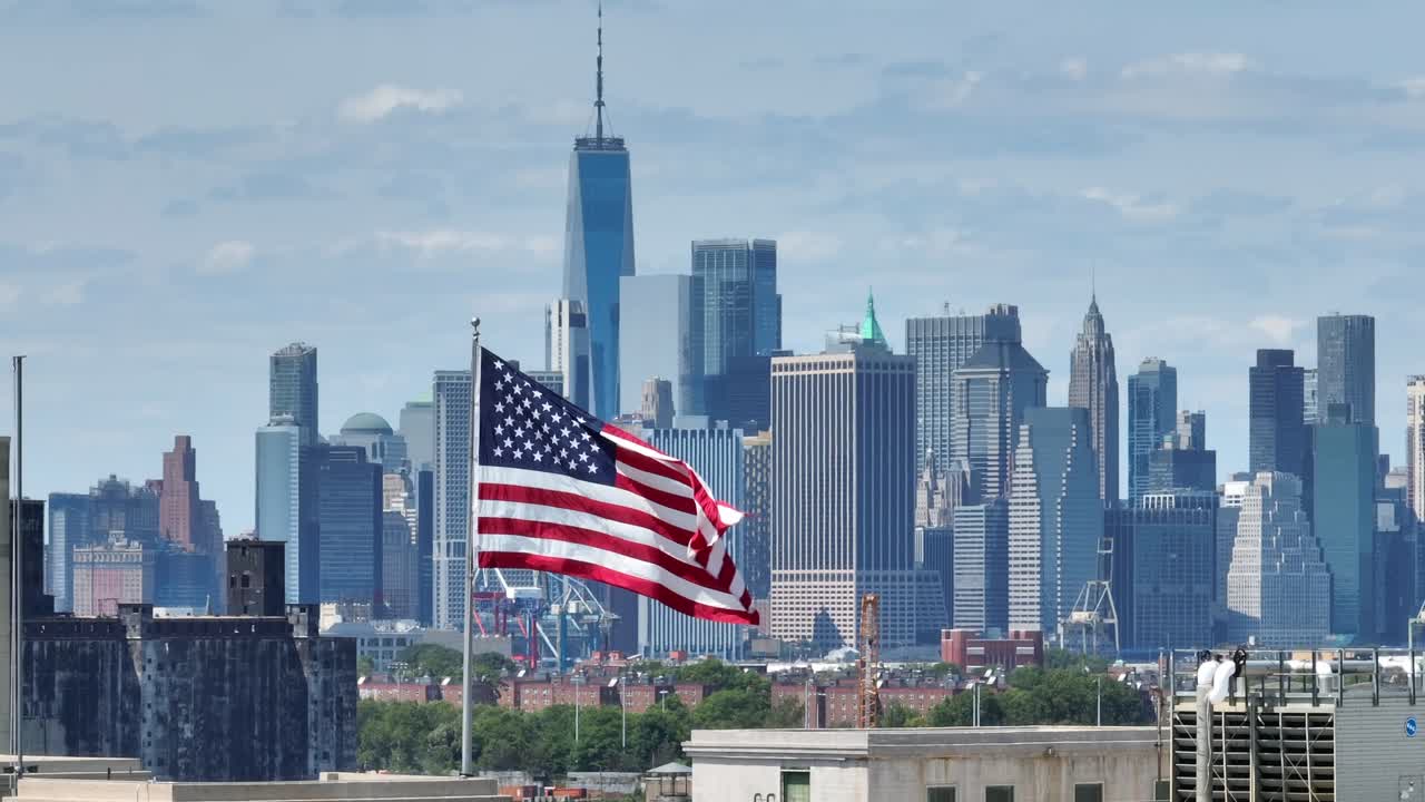 Aerial view of waving American flag in front of skyline in New York City with modern one world trade center in sunlight - Establishing drone shot view from Brooklyn District