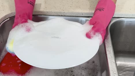 person wearing pink gloves scrubs and rinses a dirty plate under running water in a well-lit kitchen, finishing by placing it on a drying rack