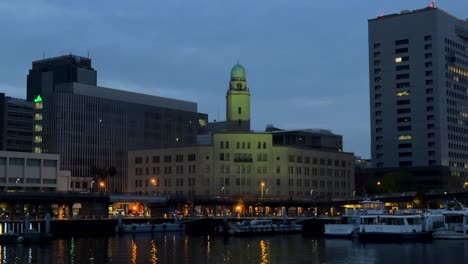 Twilight-cityscape-with-illuminated-buildings-by-the-harbor,-boats-docked,-serene-ambiance,-wide-shot