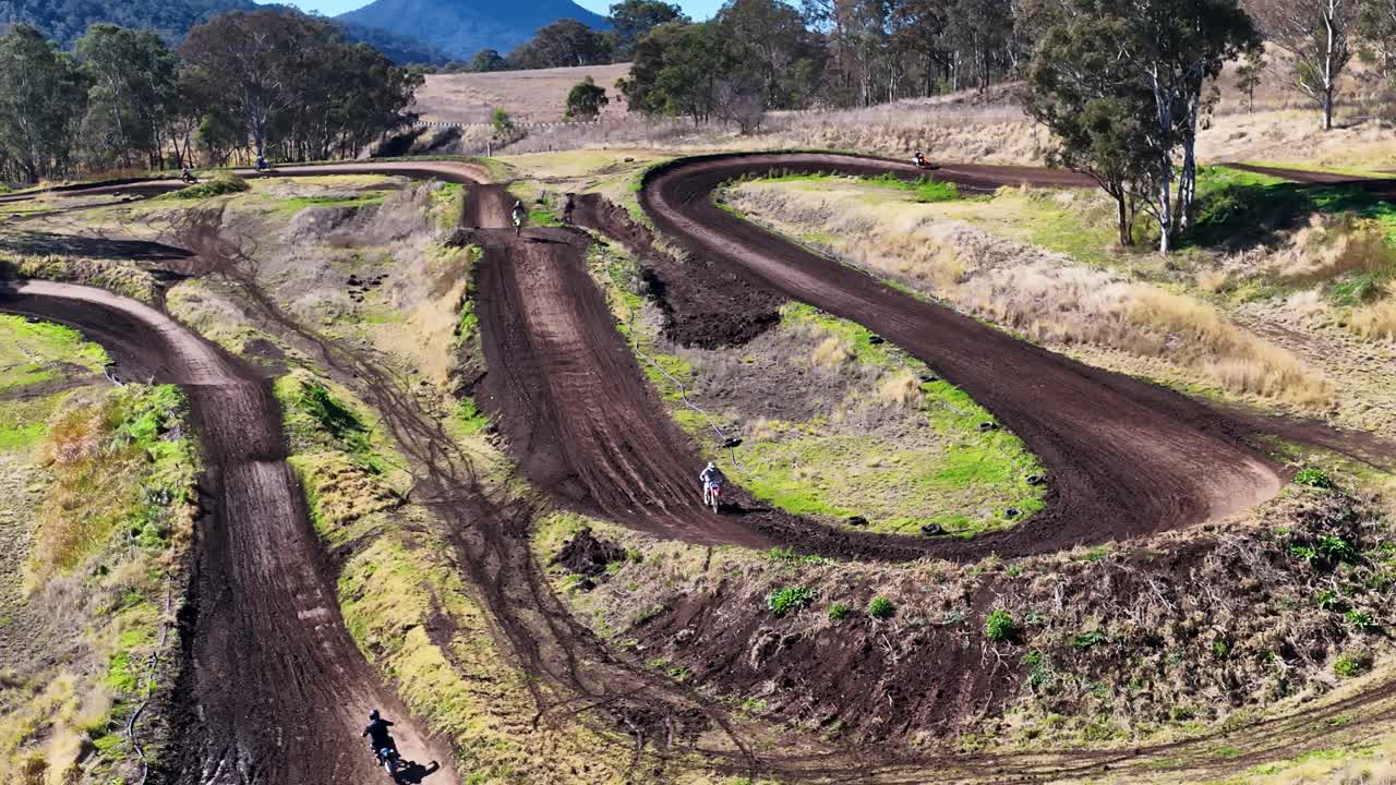 Motocross rider speeds along winding dirt track, rural landscape, bright daylight, aerial perspective