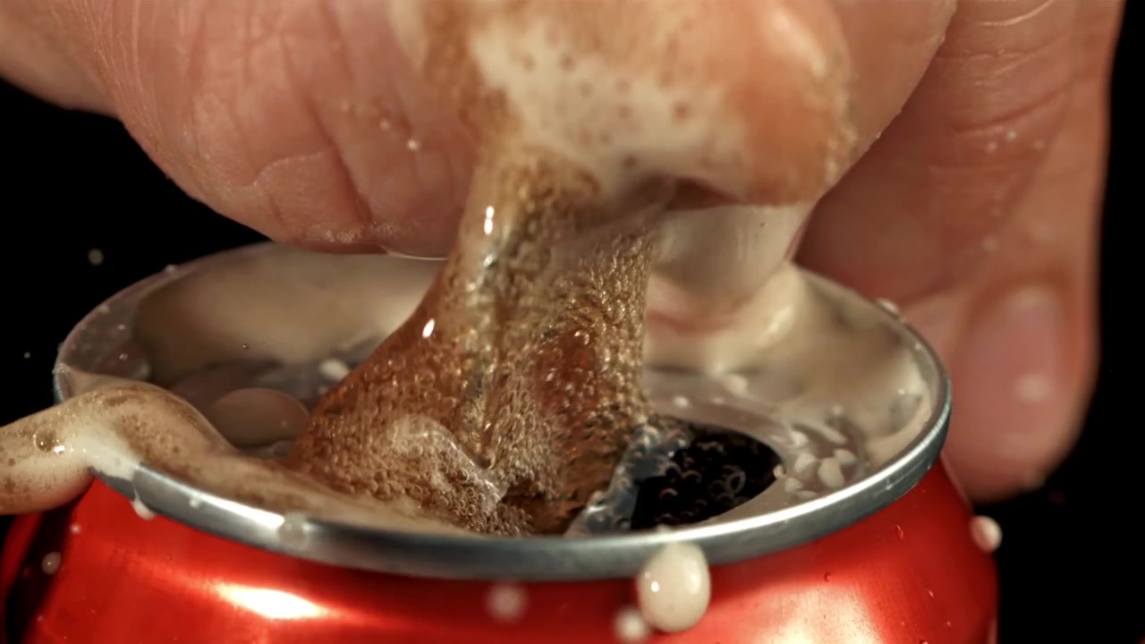 Primo piano di una mano che apre una lattina di soda rossa con un'effervescenza esplosiva.