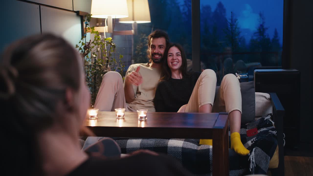 un chico y una chica están sentados en un sofá y charlan con una chica rubia en una mesa en la que hay velas. conversación acogedora de amigos por la noche