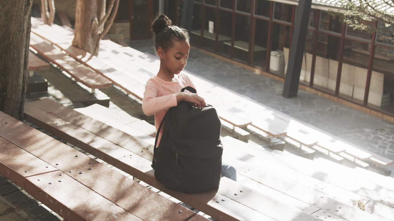 In school, African American girl sitting on bench opening backpack, preparing for class