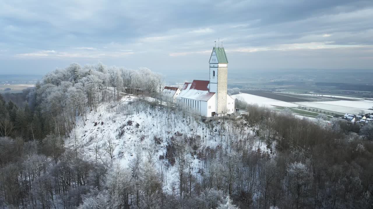 Drone footage of a snowy winter landscape as it moves away from a hilltop church, revealing a serene snow-covered forest in the foreground. A peaceful and cinematic winter scene.