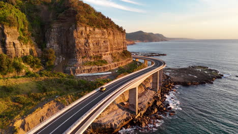 breathtaking aerial drone shot of a car driving along the sea cliff bridge, capturing the dramatic ocean cliffs and curving roadway. golden hour.