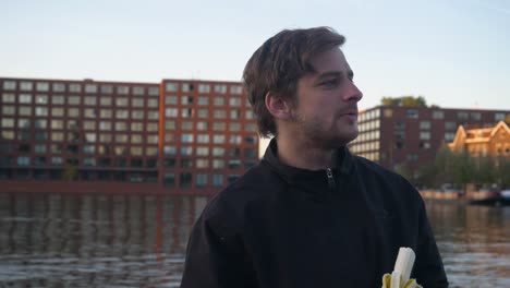 close up of a young handsome man peeling and eating a banana with an urban pier in the background, amsterdam