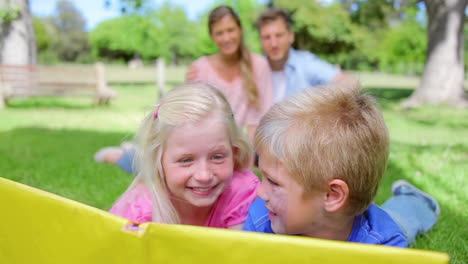 Two-kids-reading-a-book-together-while-smiling-and-looking-at-each-other-as-they-lie-on-the-grass