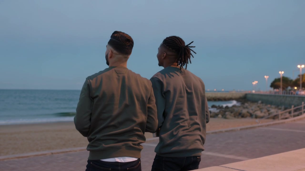 Toasting two men on low sea wall, watching waves, rising, standing on walkway holding metal bottles
