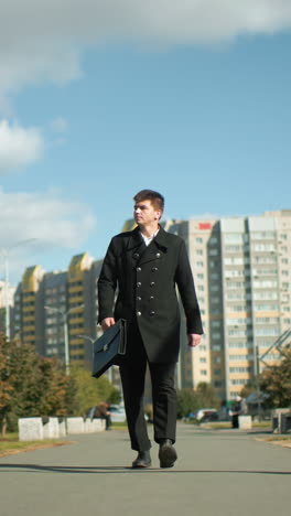 joven caminando con confianza por una carretera pavimentada con alquitrán llevando una bolsa, rodeado de árboles vibrantes, edificios residenciales modernos, coches estacionados y una figura borrosa en el fondo lejano bajo un cielo azul brillante