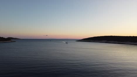 Stunning-aerial-of-a-drone-at-middle-altitude-flying-out-of-a-cove-while-a-boat-approaches-as-sun-sets-in-Croatia