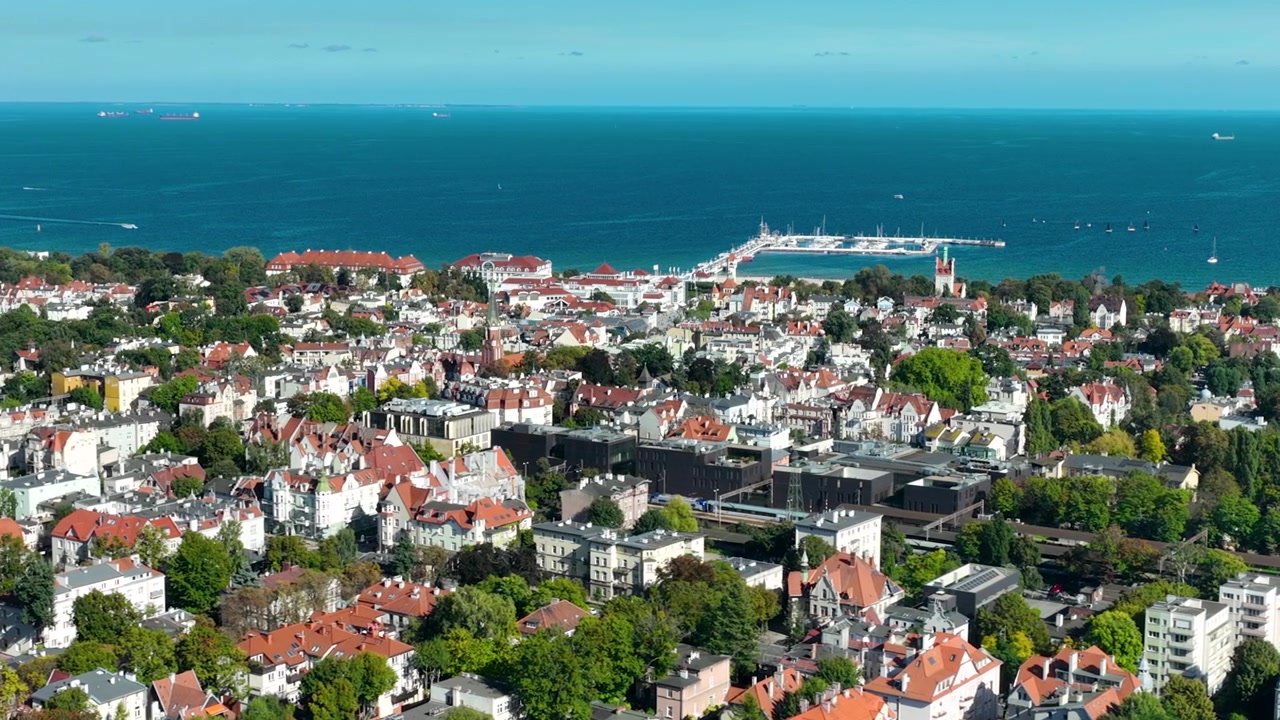 Vista aérea de ângulo alto de uma bela cidade litorânea com arquitetura de telhados vermelhos, árvores verdes exuberantes e um longo píer de madeira estendendo-se para o mar azul