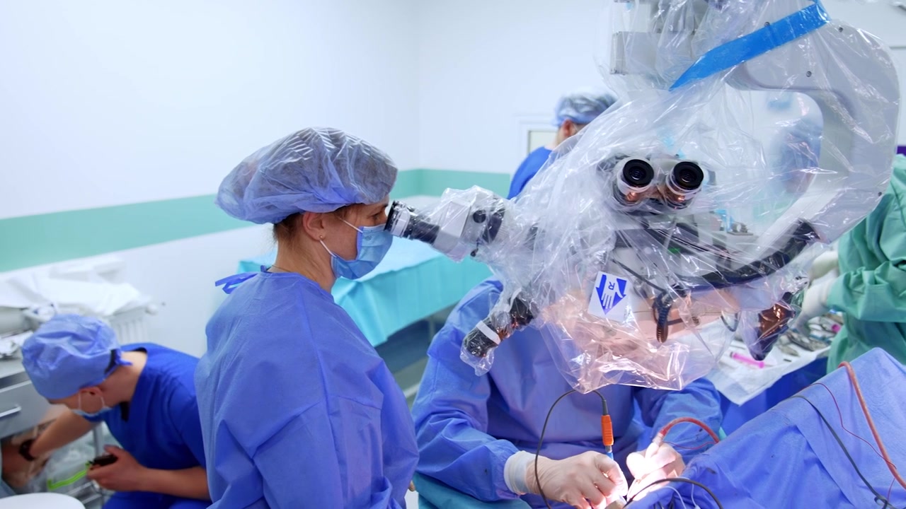 Una médica se para mirando el microscopio. Un cirujano no identificado se sienta detrás del microscopio usando instrumentos durante la operación