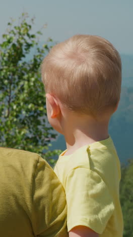 long haired blonde mother holds little son in arms and points forward at ropeway cabins against misty old forestry mountains backside view slow motion