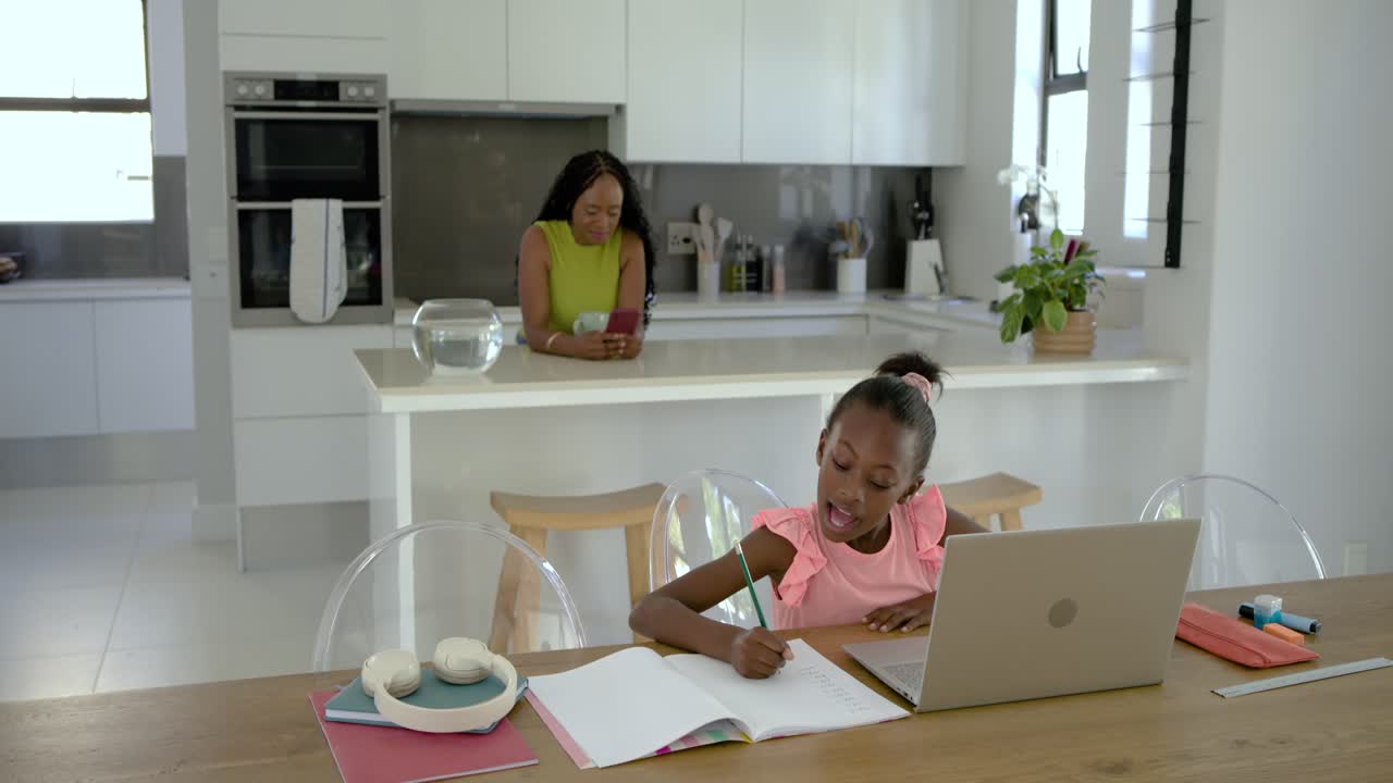 Mother and daughter in kitchen, student girl in pink waving for online class writing at laptop
