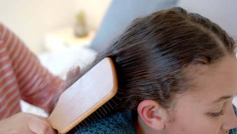 Happy-biracial-mother-brushing-daughter's-hair-in-sunny-bedroom