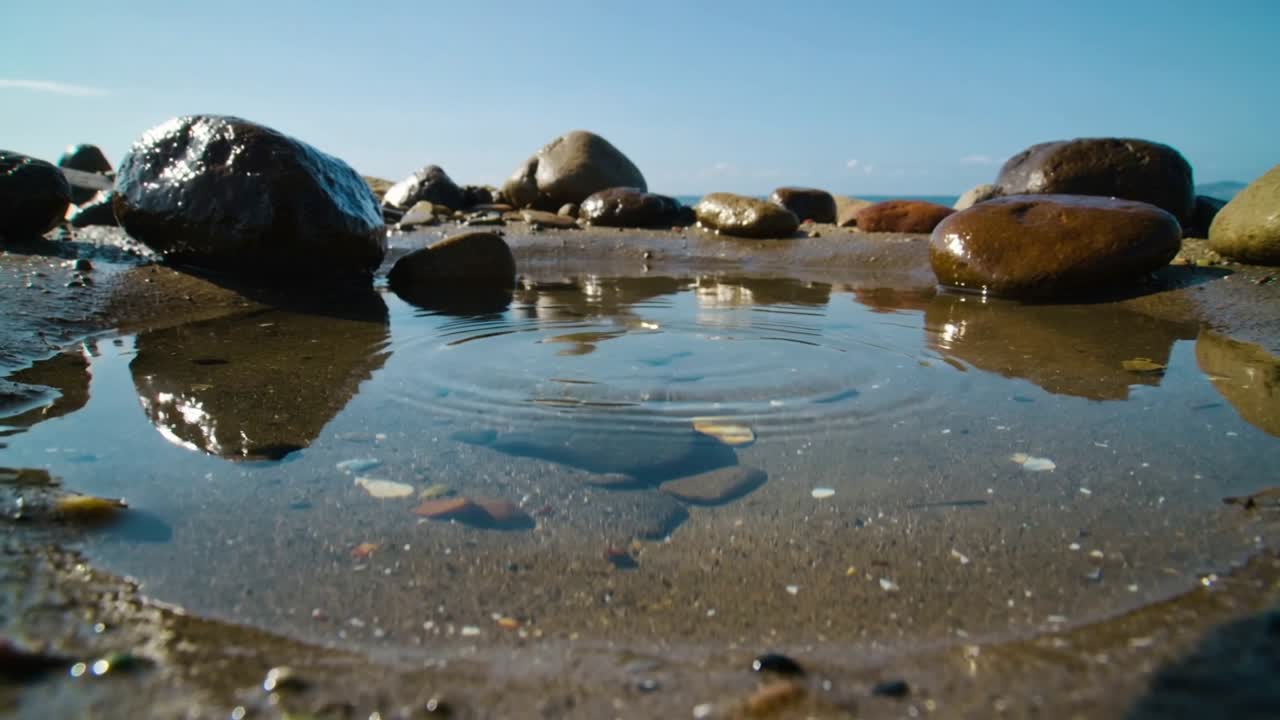 Pozza di marea increspata dopo che una piccola goccia colpisce il centro sulla sabbia, mostrando ciottoli e il riflesso del cielo.