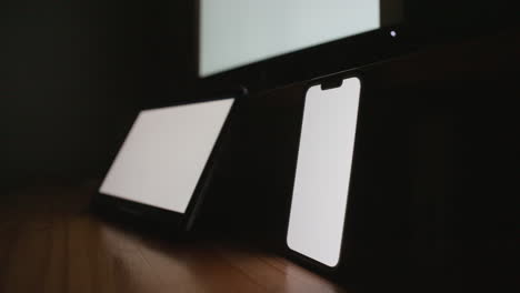 Three-blank-monitors-and-devices-on-a-desktop-in-a-dark-room-with-a-white-luma-screen-displayed-for-compositing-purposes