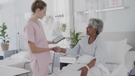 happy diverse female doctor using tablet talking with senior female patient in bed, slow motion