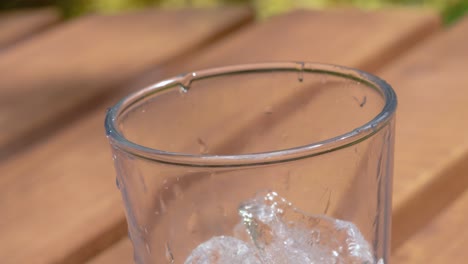 Melting-Ice-Cubes-Falling-into-Clear-Glass-Sitting-on-Picnic-Table-on-Sunny-Summer-Day---Ultra-Slow-Motion-4K