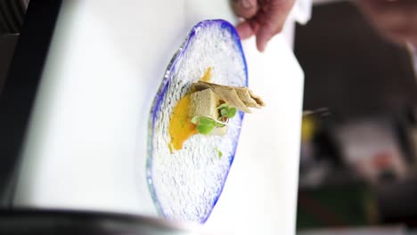 Vertical-slow-motion-shot-of-a-gourmet-meal-of-tofu-garnished-with-green-leaves-doused-with-olive-oil-in-an-upscale-restaurant