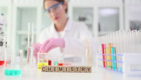 A person in a lab coat and safety glasses working with colorful liquids and lab equipment, with the word 'CHEMISTRY' spelled out in front