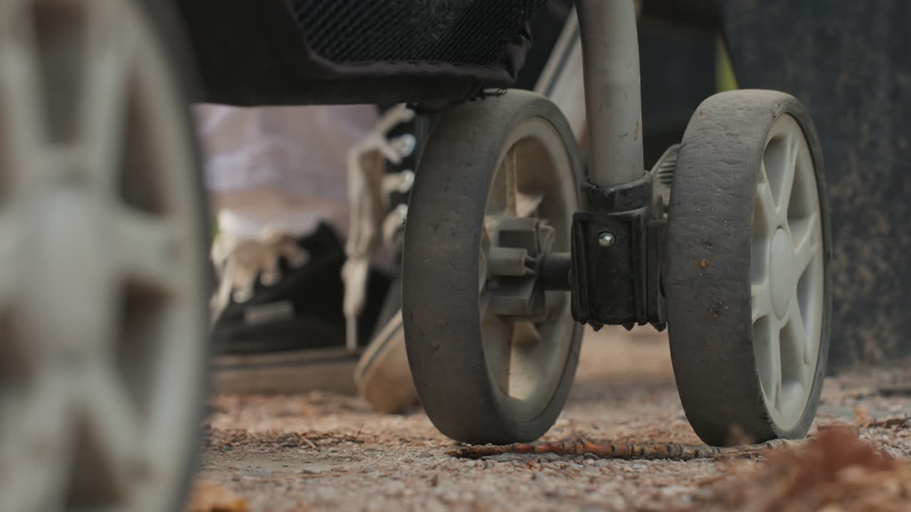 Lowangle stroller wheels on pavement, city sidewalk at dusk with worn treads and textured asphalt, sneakered parent moving steady pace, cinematic moody tones, reflective family routine during quiet