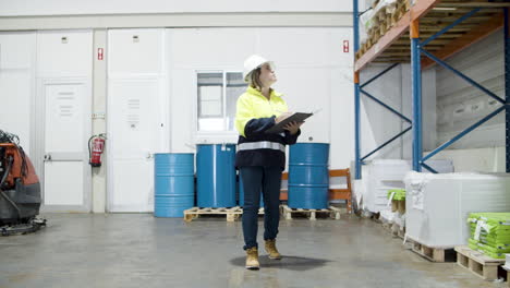 serious caucasian female worker in helmet holding clipboard and counting goods in stock