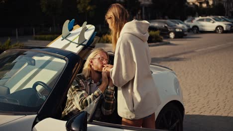 A-blonde-girl-in-a-White-sweatshirt-stands-near-her-blonde-boyfriend-in-a-checkered-shirt-who-sits-in-a-White-convertible-and-kisses-his-girlfriend’s-hand.-Happy-couple-near-their-Convertible-with-surfboards-in-the-city