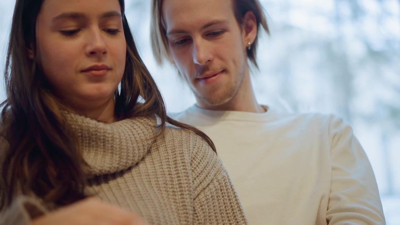 Primo piano di due persone in piedi insieme, una donna con un maglione lavorato a maglia e un uomo accanto a lei. La scena è illuminata delicatamente da luce naturale che filtra attraverso un vetro smerigliato, creando un'atmosfera interna intima.
