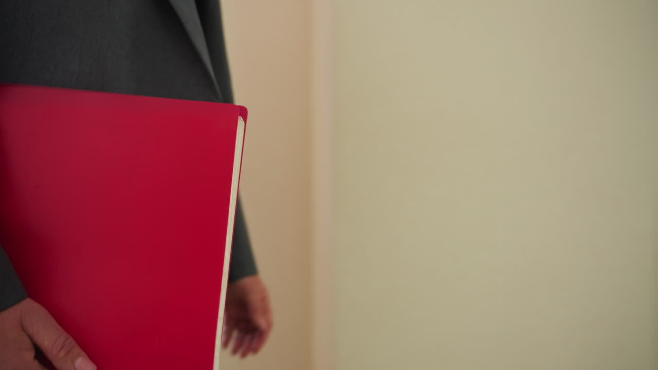 Professional person dressed in gray corporate outfit walking indoors while holding bright red envelope under arm in modern office hallway with sunlight filtering through window in background