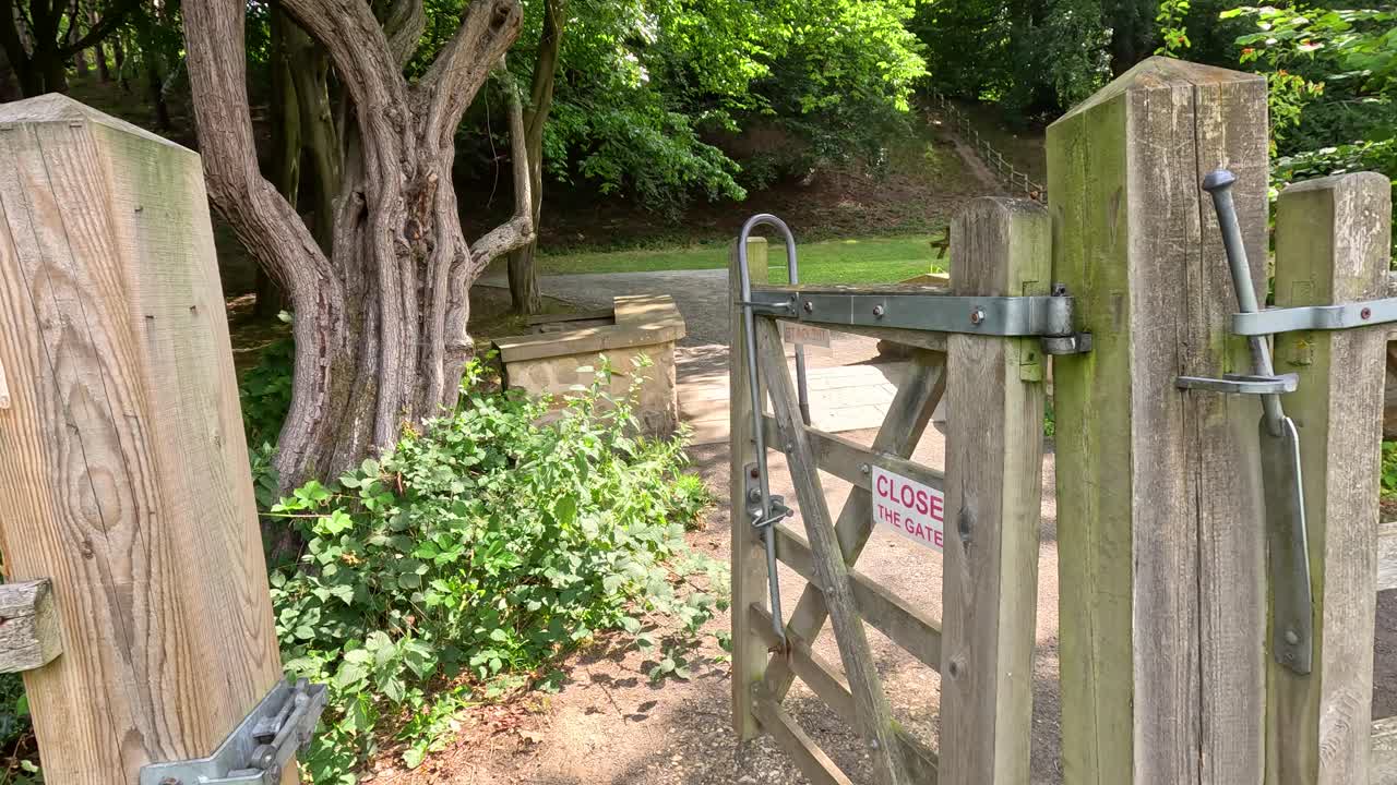 A wooden gate is manually closed along a leafy garden path in daylight, with a visible sign requesting closure. Natural lighting, steady camera, tranquil mood