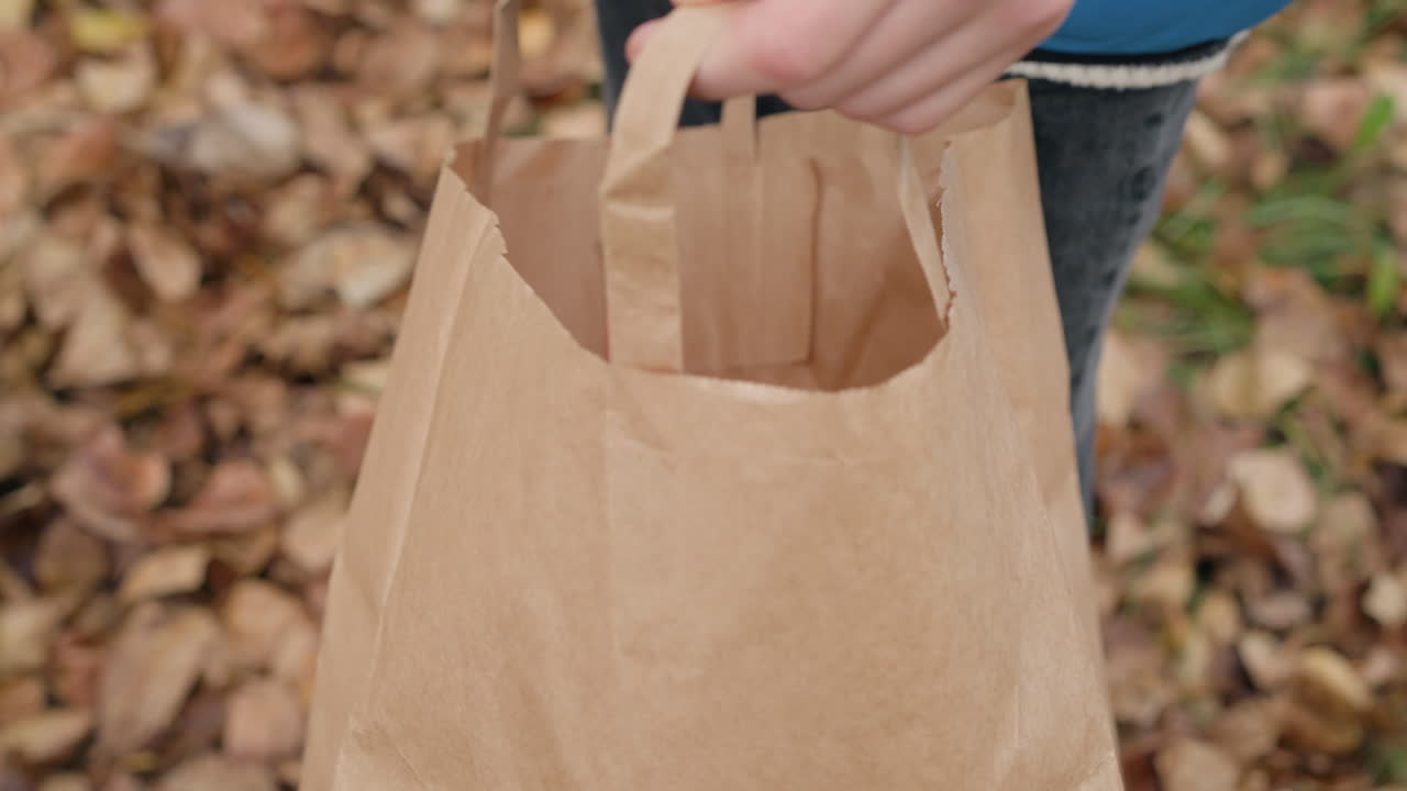 primer plano de un niño colocando una hoja seca de otoño en una bolsa de papel marrón, mostrando una actividad al aire libre sostenible e interacción con la naturaleza rodeada de hojas caídas