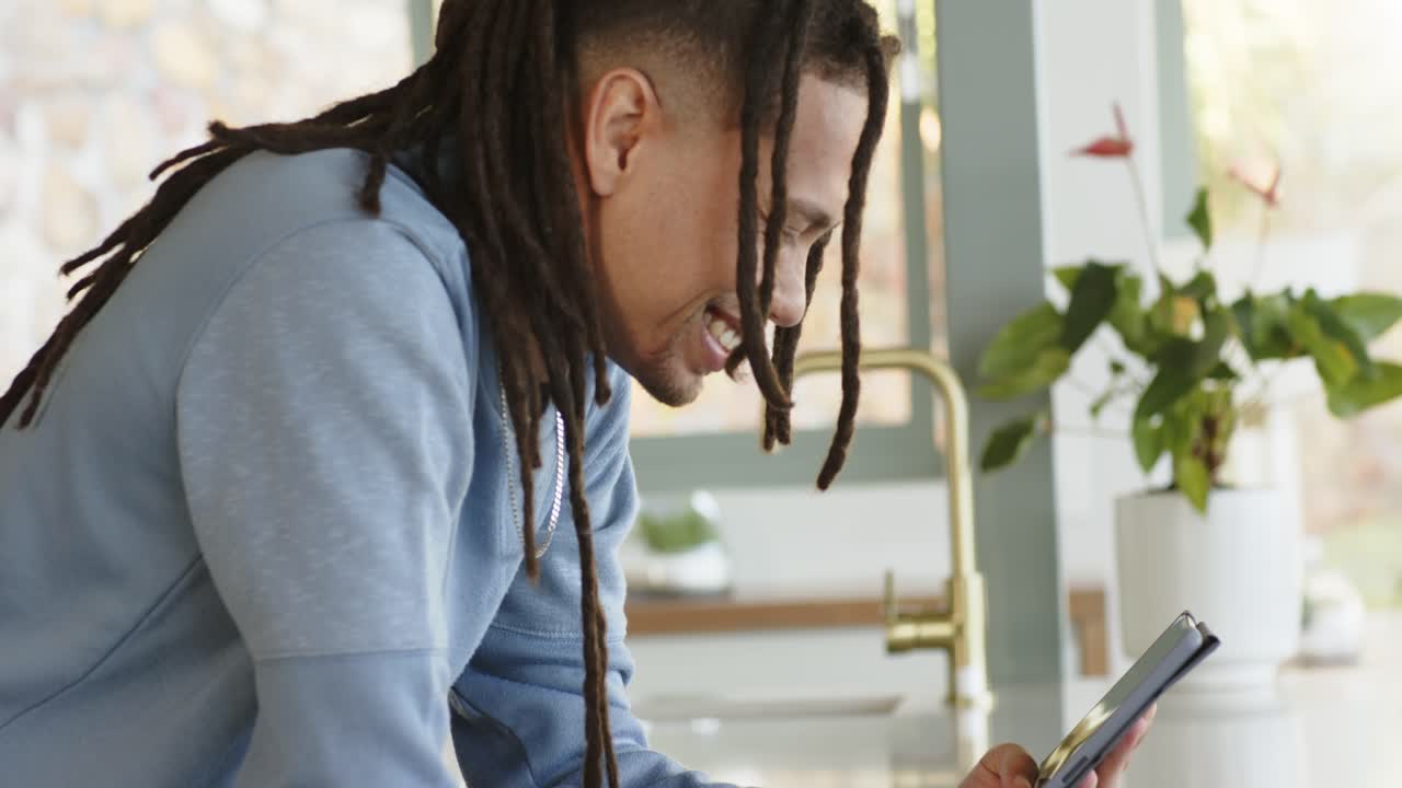 Middle aged man with dreadlocks leaning on kitchen island reading smartphone tapping while smiling