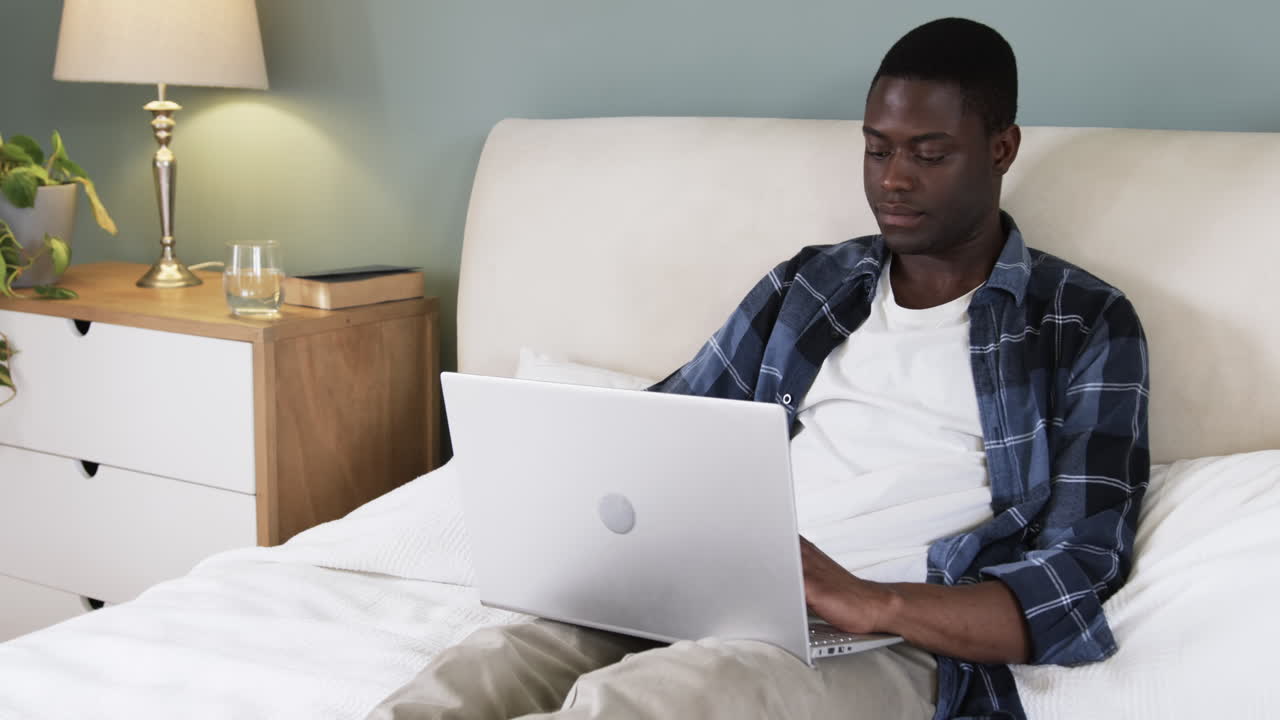 African American man using laptop on bed at home, focusing on work