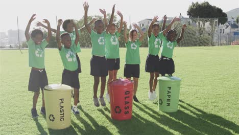 Portrait-of-happy-diverse-schoolchildren-with-recycling-bins-in-sports-field-at-elementary-school