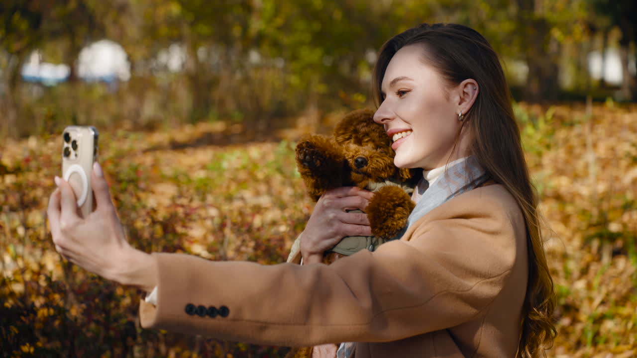A woman is smiling while taking a selfie outdoors, holding a small Toy Poodle brown dog, with autumn leaves in the background - close-up parallax