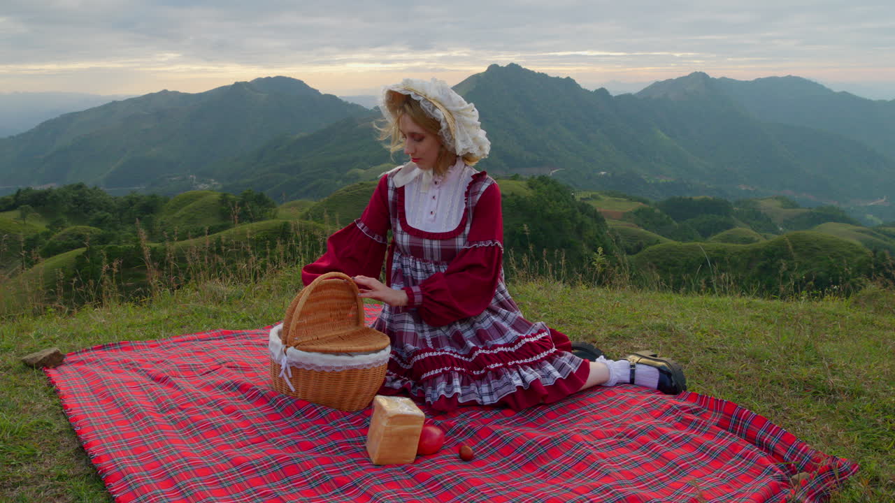 빨간색 플레이드 드레스를 입은 예쁜 여자가 피크닉 담요에 앉아서 산 ⁇ 에서 바구니에서 과일과 샌드위치 물건을 끌고 나갑니다.