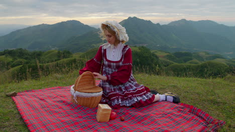 mujer bonita en vestido a cuadros rojo se sienta en una manta de picnic tirando de frutas y sandwhich artículos de la cesta en la ladera vista