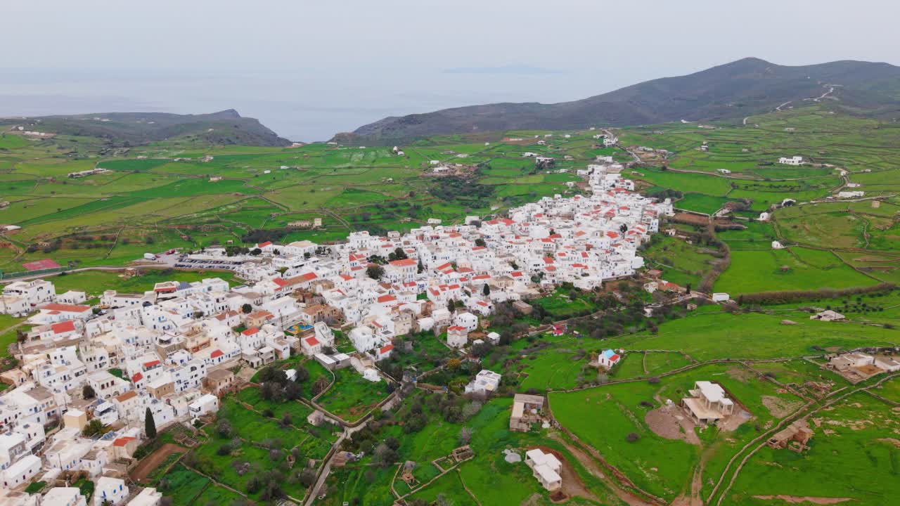 Wide drone overview of Kythnos town nestled in valley with open green terrain and scattered buildings, backdrop background
