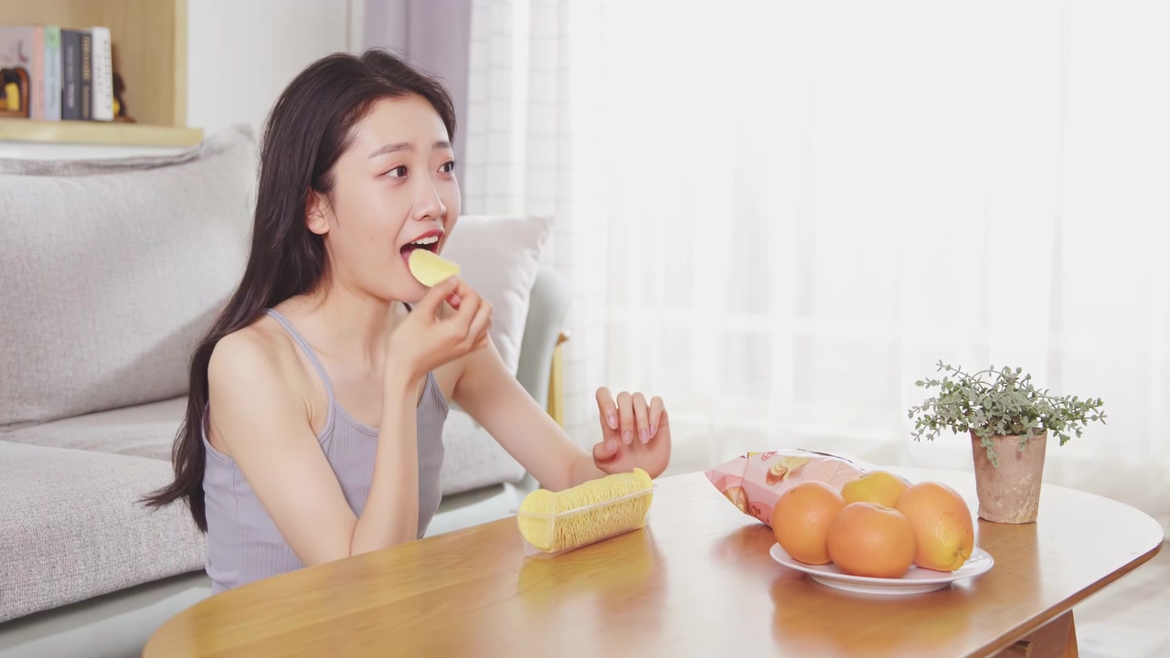 아름답고 매력적인 젊은 여성이 집 거실 소파에 앉아 봉지에서 바삭한 감자칩을 즐겁게 먹고 있습니다. 휴식을 취하며 TV 리모컨을 켭니다.