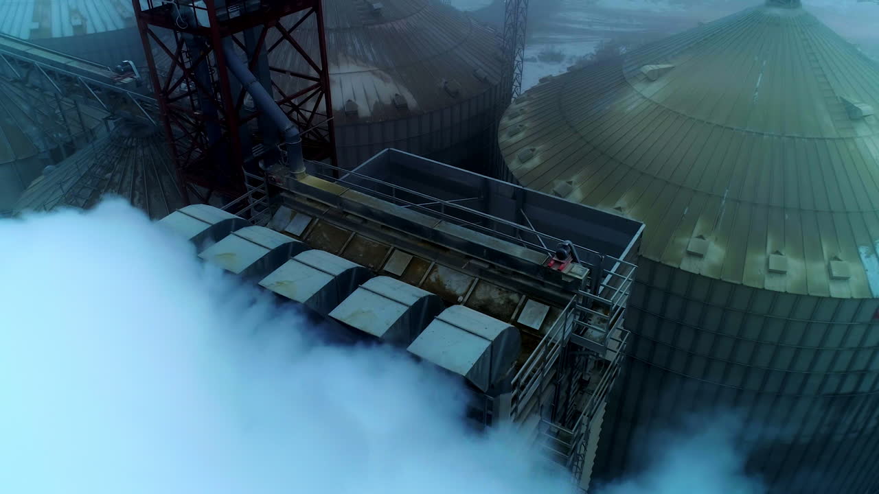 Five thick pipes letting out heavy white smoke as a result of grain processing. Rising over the big agricultural plant at foggy weather day.