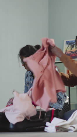 excited ladies throw clothes into suitcase sitting on floor near sofa. african american and indian women prepare for summer holiday abroad slow motion