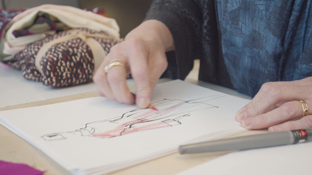 An older woman sketching a woman in a dress, with colored pencils, fabric swatches, and other fashion drawings arranged on her workspace