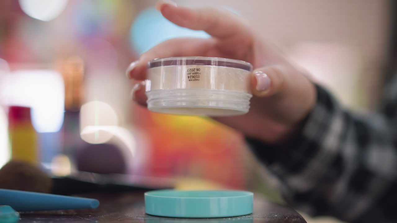 primer plano de la mano de la joven tirando polvo en la cubierta, preparando maquillaje, artículos de maquillaje de colores y pinceles visibles en la mesa, indicando la rutina de belleza y el proceso de aplicación de maquillajes