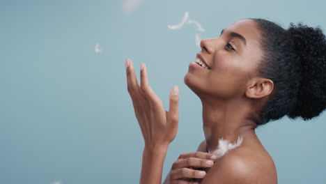 slow motion portrait beautiful african american woman feathers falling on smooth skin touching bare shoulders enjoying natural skincare beauty gently caressing body in blue background