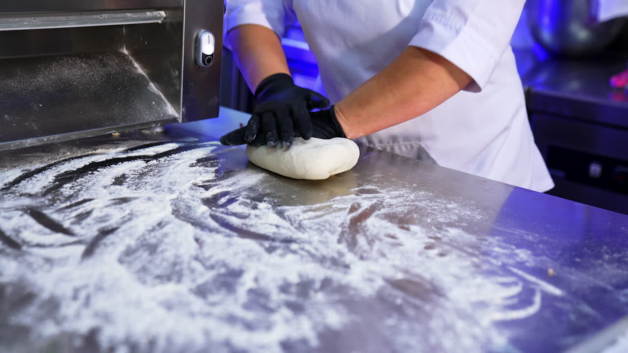 Culinary specialist in black latex gloves kneading the dough. The cook prepares dough on a big metal table covered with flour.