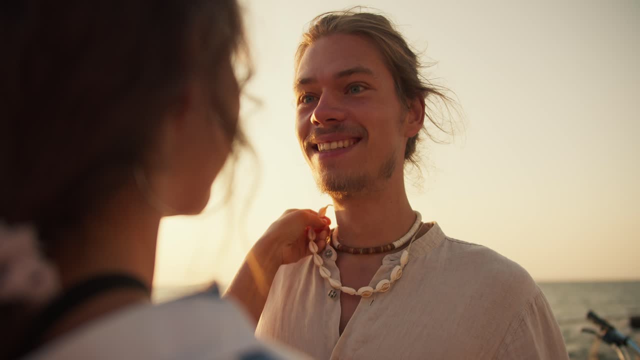 Close-up shot of a girl in a white blue shirt stroking her boyfriend on the cheek and adjusting his jewelry on his neck near the sea at Sunrise in summer. Happy guy with stubble wearing white clothes and a white necklace