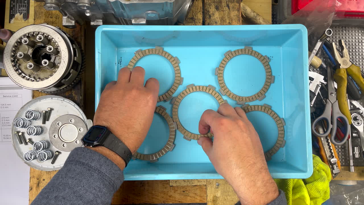 Close-up overhead POV shot of a mechanic placing five new kevlar clutch plates into a clean blue plastic basin, ready for oil soaking during DIY scooter engine maintenance