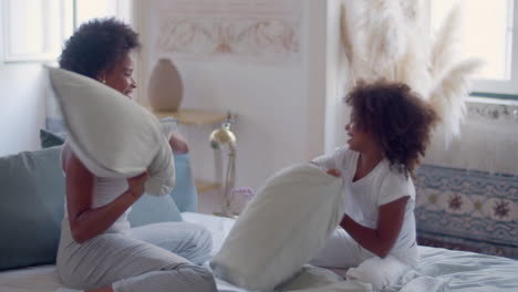 happy african american mum and daughter having pillow fight in bed