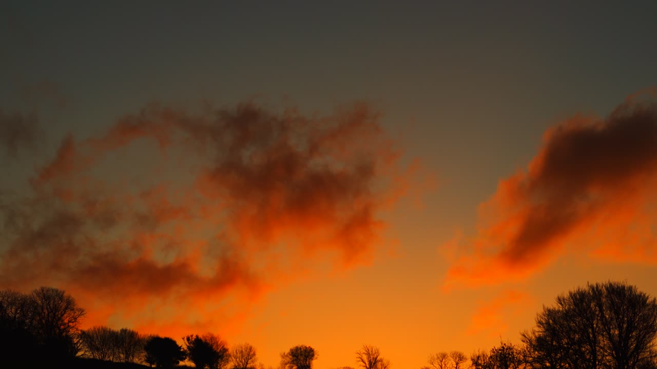 Orange Sunset Clouds Slowly Moving Across Sky and Sun Sets Beyond Horizon with Silhouetted Natural Trees. Rural United Kingdom. Beautiful Nature Footage 4K